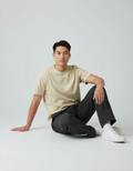 Man sitting on the floor wearing a beige t-shirt and black pants against a white background