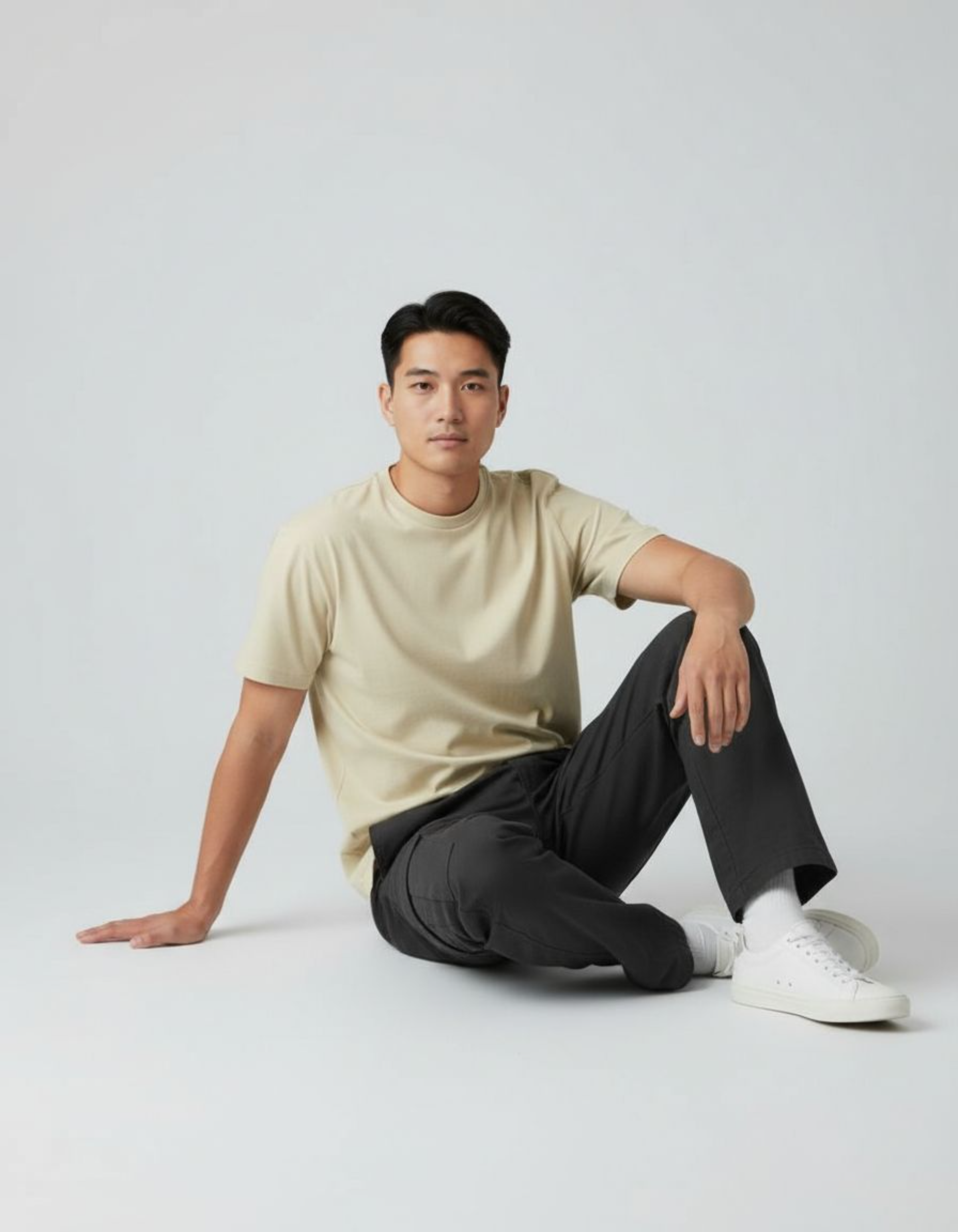Man sitting on the floor wearing a beige t-shirt and black pants against a white background