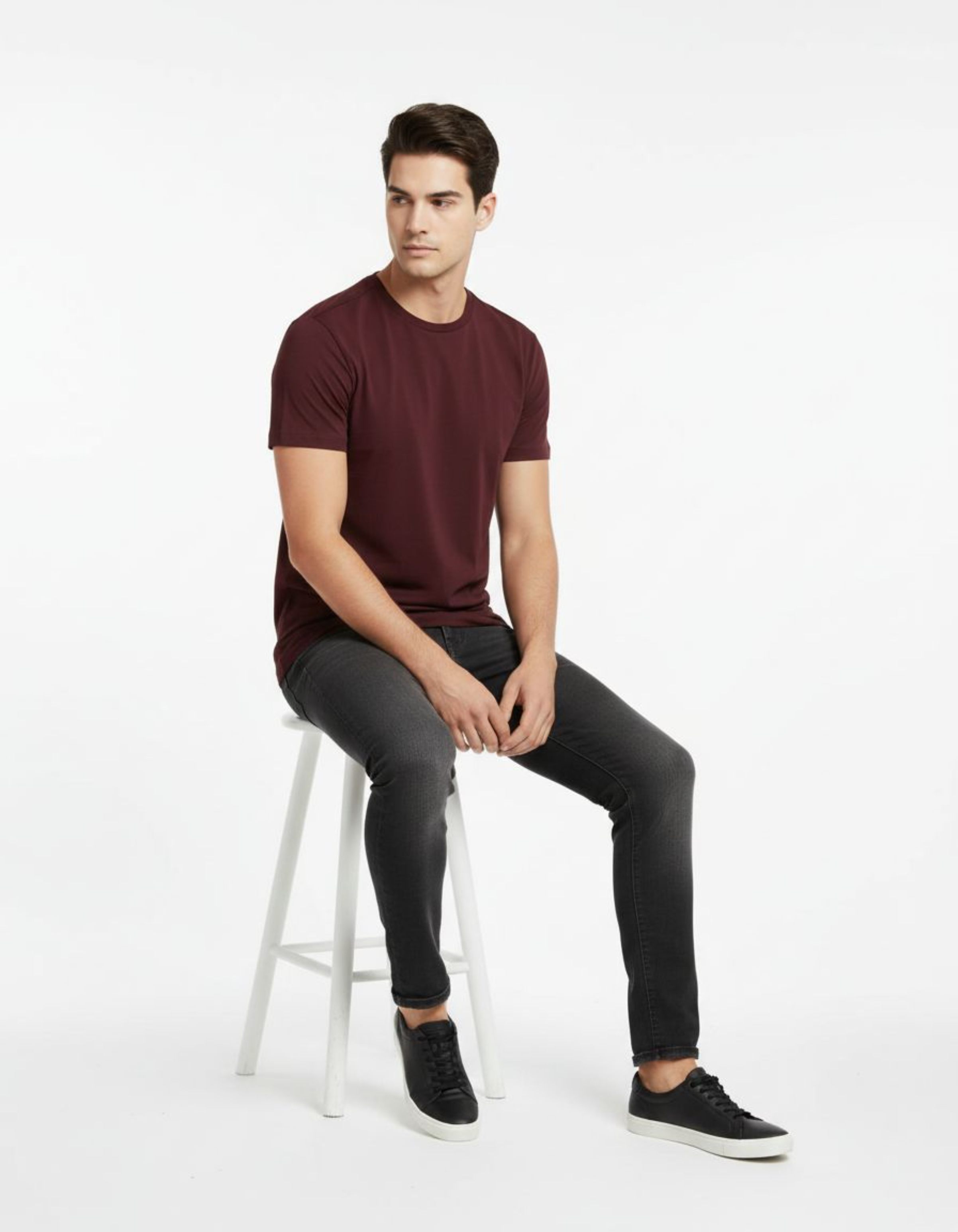 Man sitting on a white stool wearing a maroon t-shirt and black pants against a white background